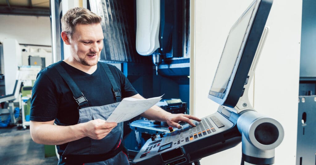 Traditional vs. CNC Machining: What’s the Difference?
A factory worker holding a piece of paper standing in front of a computer next to a CNC machine and pressing buttons.