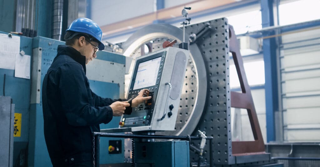 Traditional vs. CNC Machining: What’s the Difference? A man wearing a helmet and a uniform that is standing in front of a computer near a CNC machine and pressing buttons.