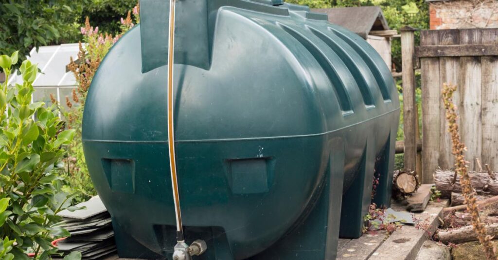 Large dark green plastic fuel tank with level gauge and valve sits on a concrete pad beside a wooden fence.