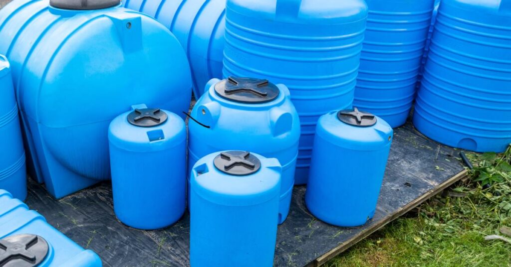 Several large and small blue storage tanks clustered together, each topped with a black cap, in a yard.