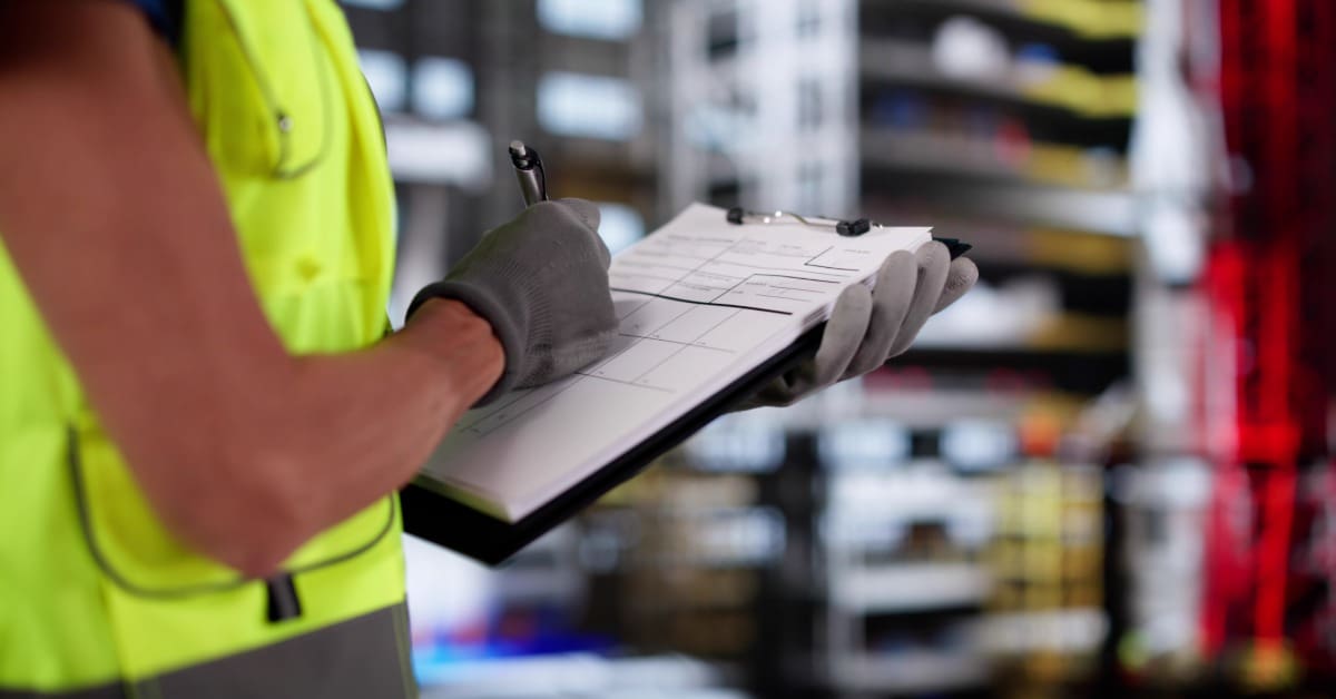 Close-up of gloved hands holding clipboard, marking checklist while standing in storage or warehouse area.