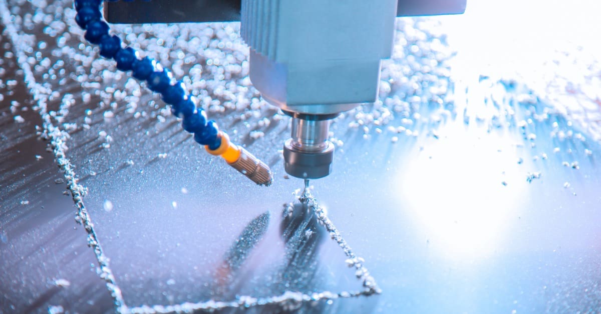 CNC milling head engraves rectangular pocket in metal plate, coolant spraying and chips scattering across surface.