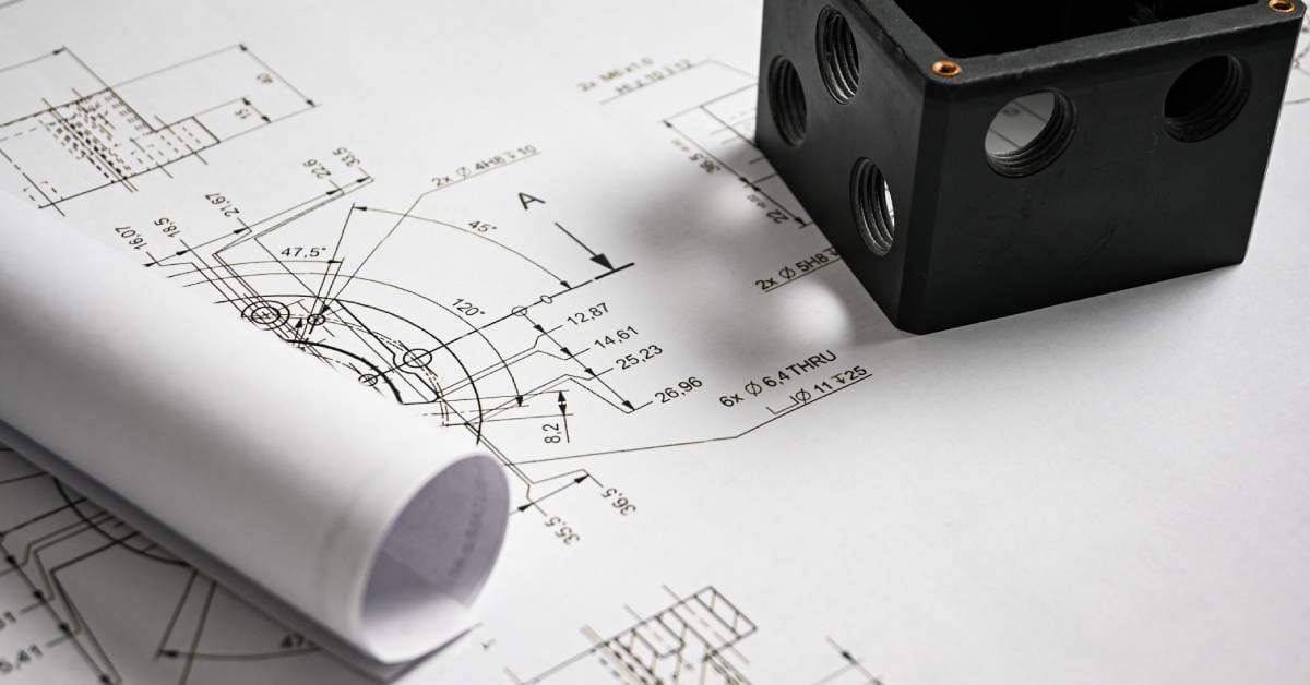 Black machined metal cube with threaded holes rests on detailed engineering drawings with measurements and diagrams.