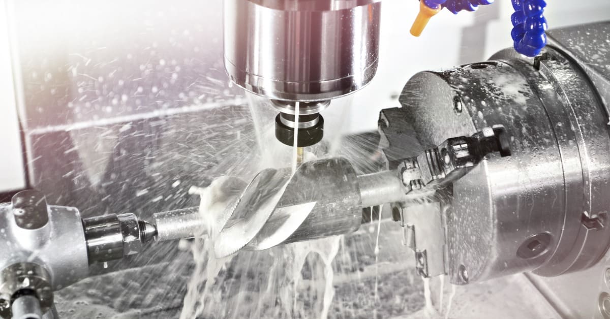 Metalworking machine shapes a large drill bit held in a rotating chuck as coolant sprays over the cutting area.