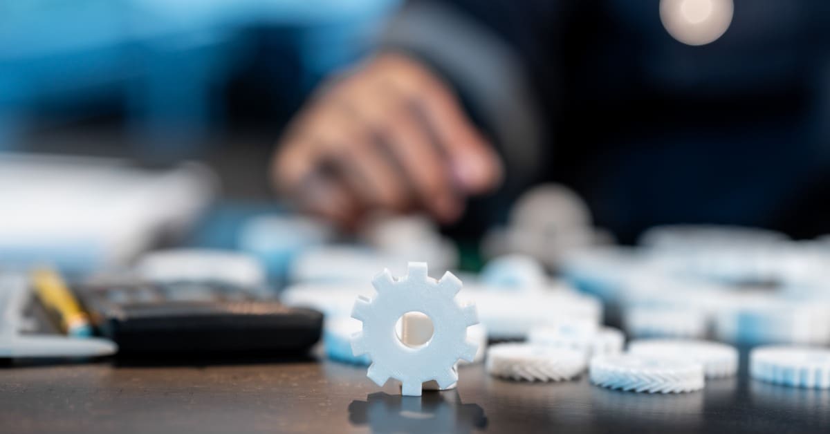 Small white gear stands upright among scattered plastic gears on a worktable, with a person blurred behind.